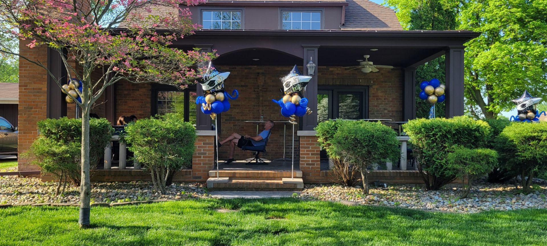 A man is sitting on a porch of a house decorated with blue and gold balloons.