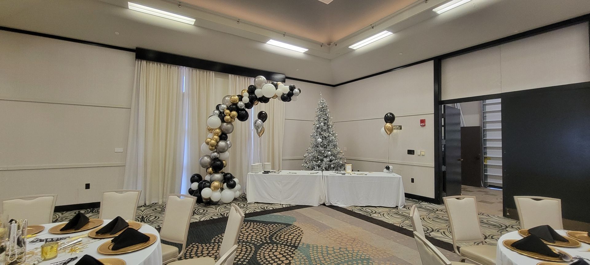 A large room with tables and chairs and a christmas tree.  Balloon demi arch