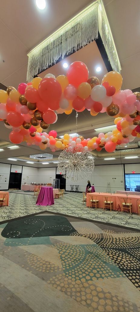 A circular balloon arch with orange, pink, and gold balloons hangs in a large room.