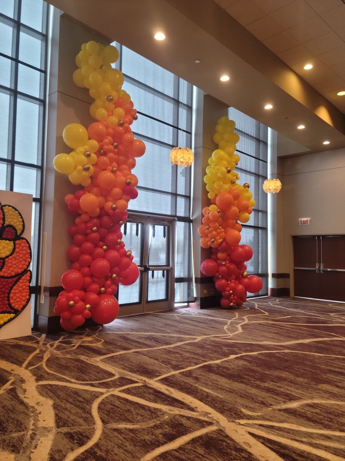 Two balloon columns in red to yellow gradient flank a doorway inside a building with large windows.