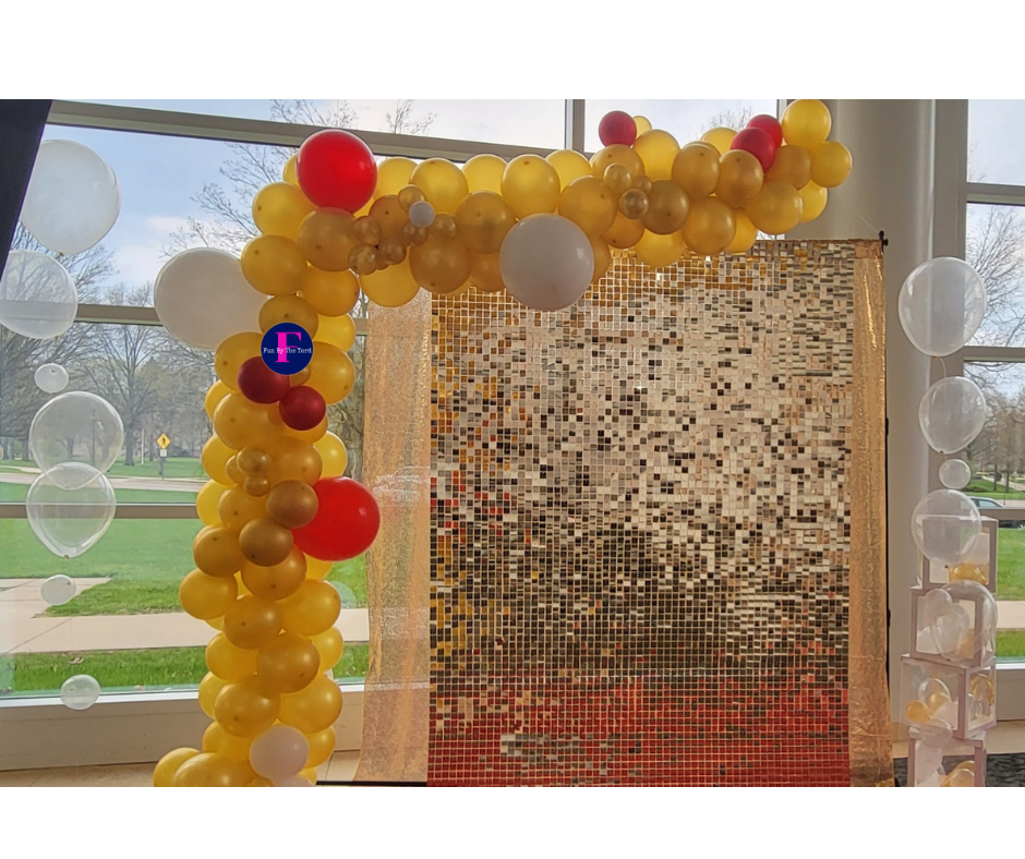 Yellow and red balloon arch and column next to a sequined gold backdrop. White balloons float nearby.