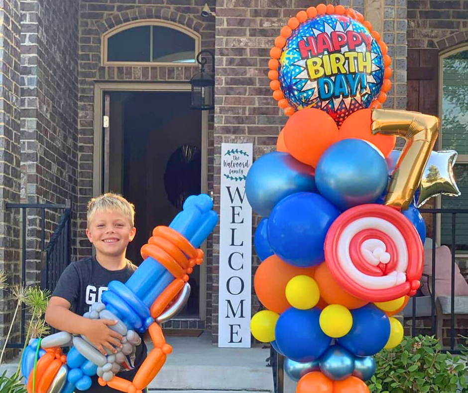 Boy holding balloon gun, smiling, next to birthday balloon display saying
