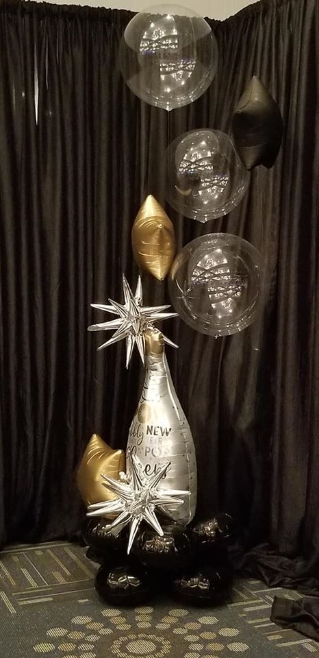 A bottle of champagne is surrounded by balloons in front of a black curtain.