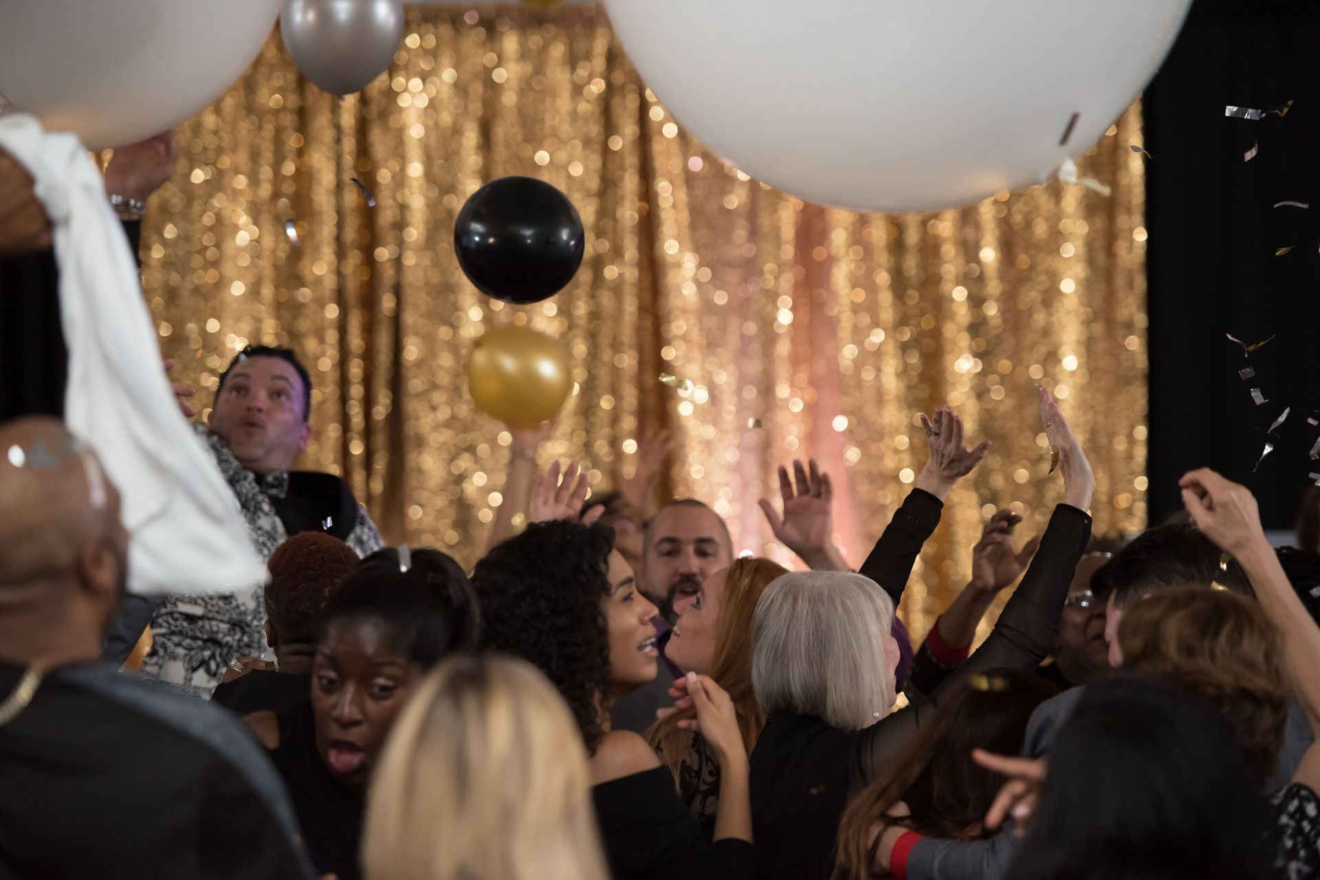 A crowd of people are dancing at a party with balloons and confetti.  Balloon drop