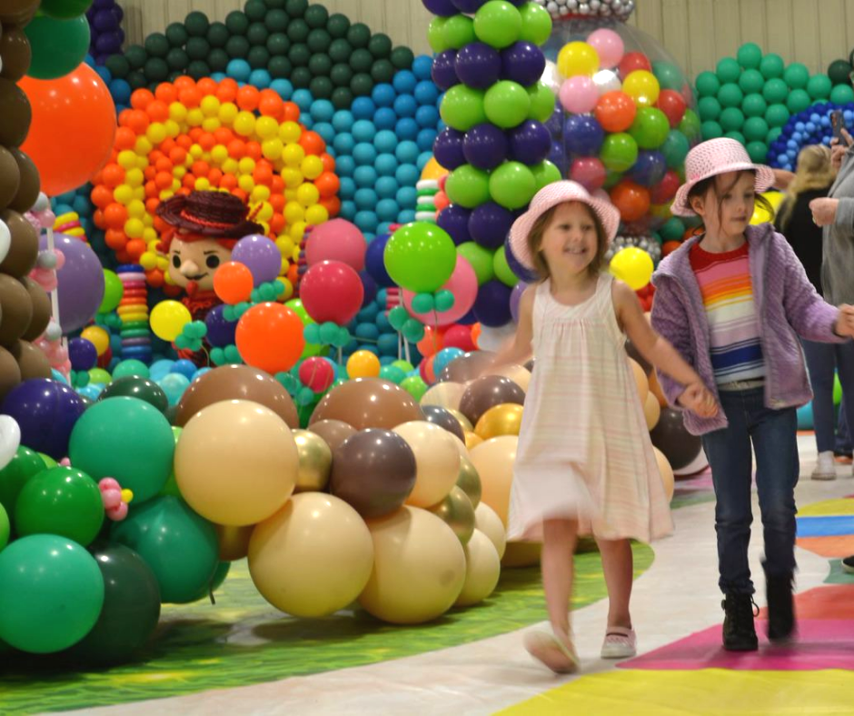 Two little girls are walking in front of a wall of balloons.  Candy Land