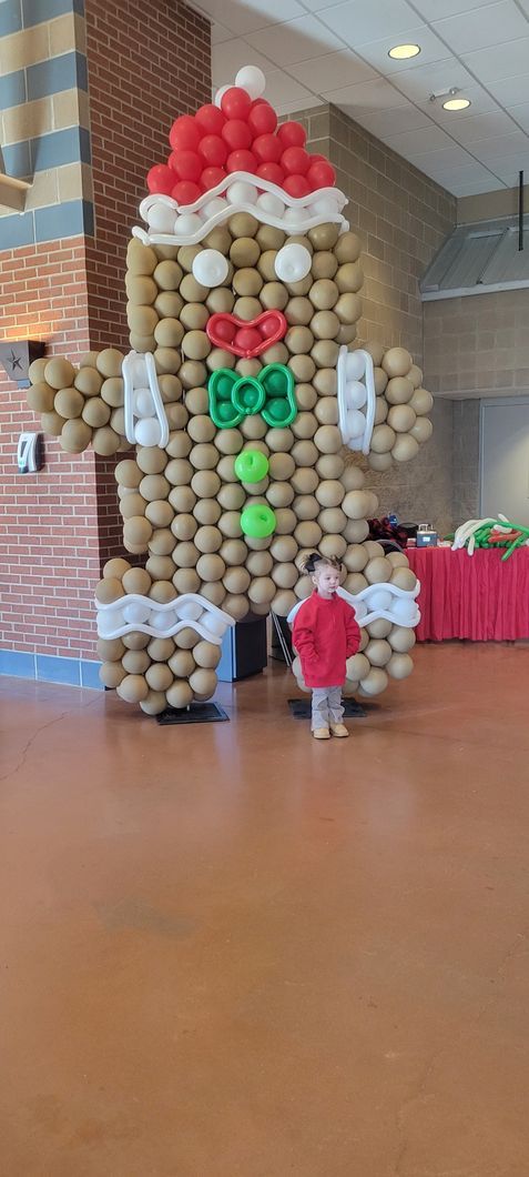 A large gingerbread man made of balloons next to a child, both indoors.