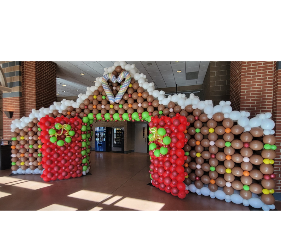 Balloon archway resembling a gingerbread house with red, brown, white, and green balloons inside a building.