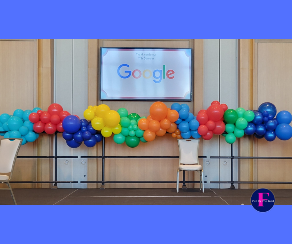 A row of colorful balloons are hanging from a wall in front of a google sign. Fun By The Yard
