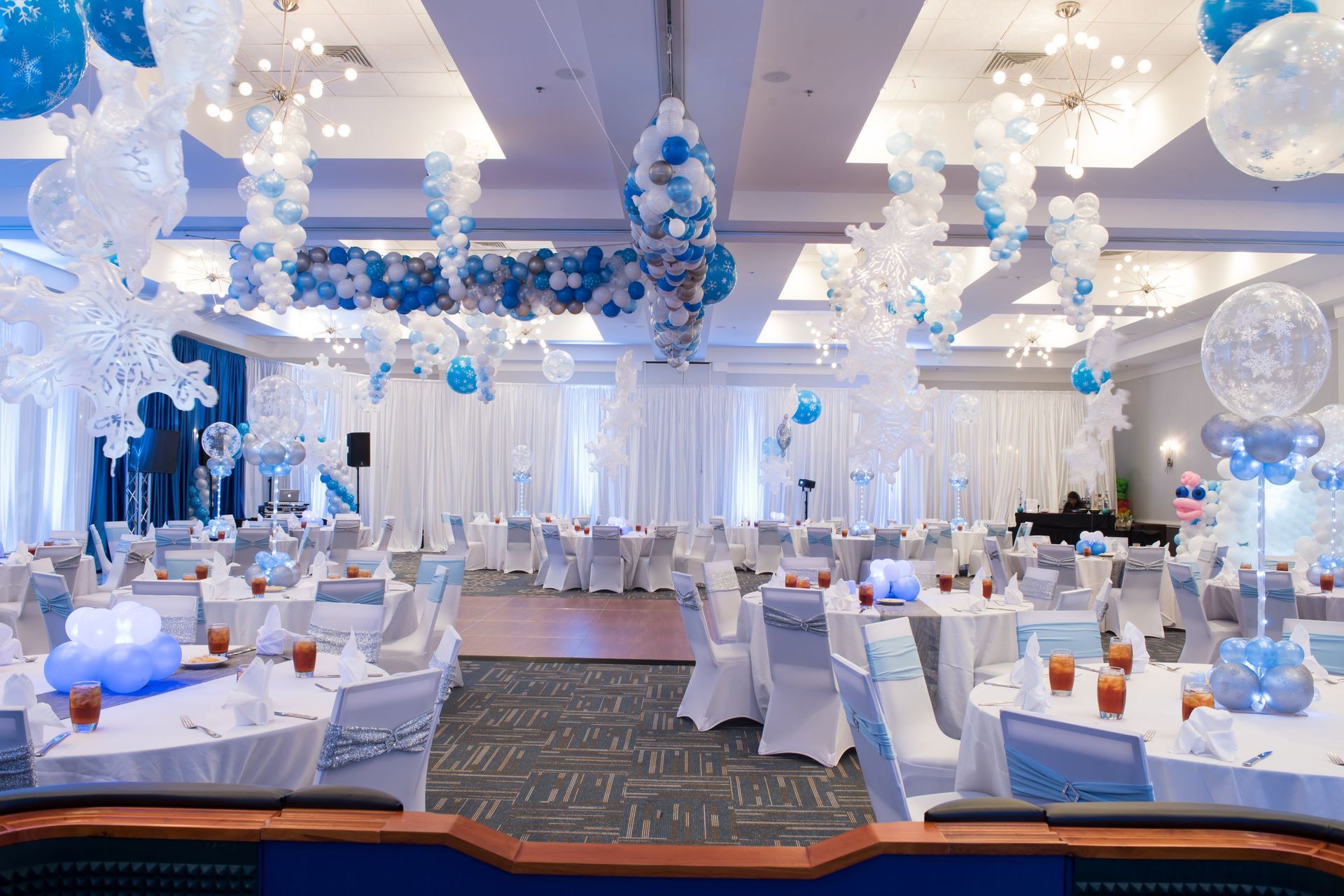 Banquet hall decorated in white and blue for a winter-themed event; tables set, balloon decorations, and stage.