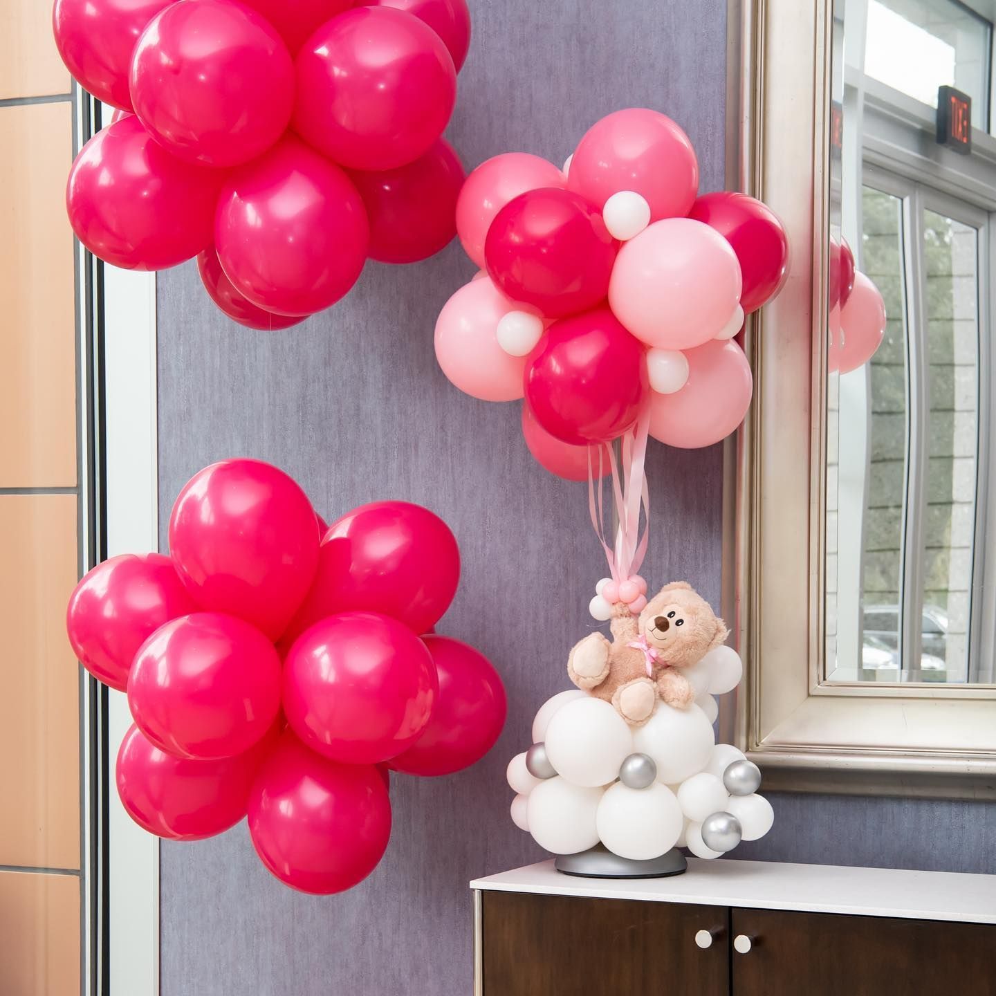 Pink and white balloon decorations, including a teddy bear arrangement, against a wall.
