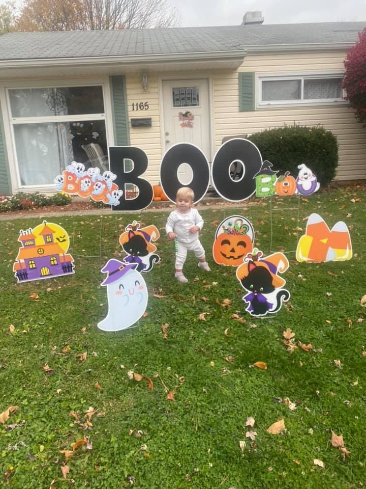 A baby is standing in front of a house decorated for halloween.  Boo themed yard card.