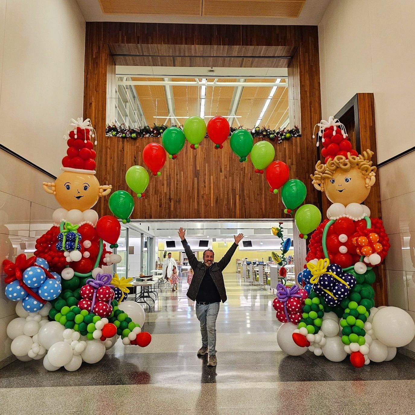 Person stands under a balloon arch in a doorway, framed by elf sculptures and gifts.