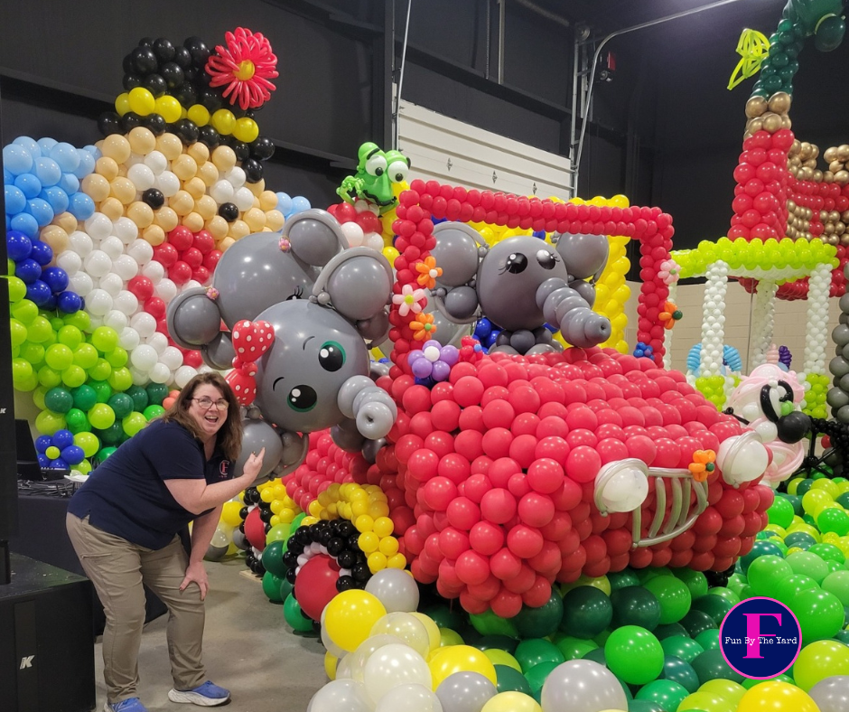 A woman is standing in front of a car of elephants made out of balloons.  Fun By The yard