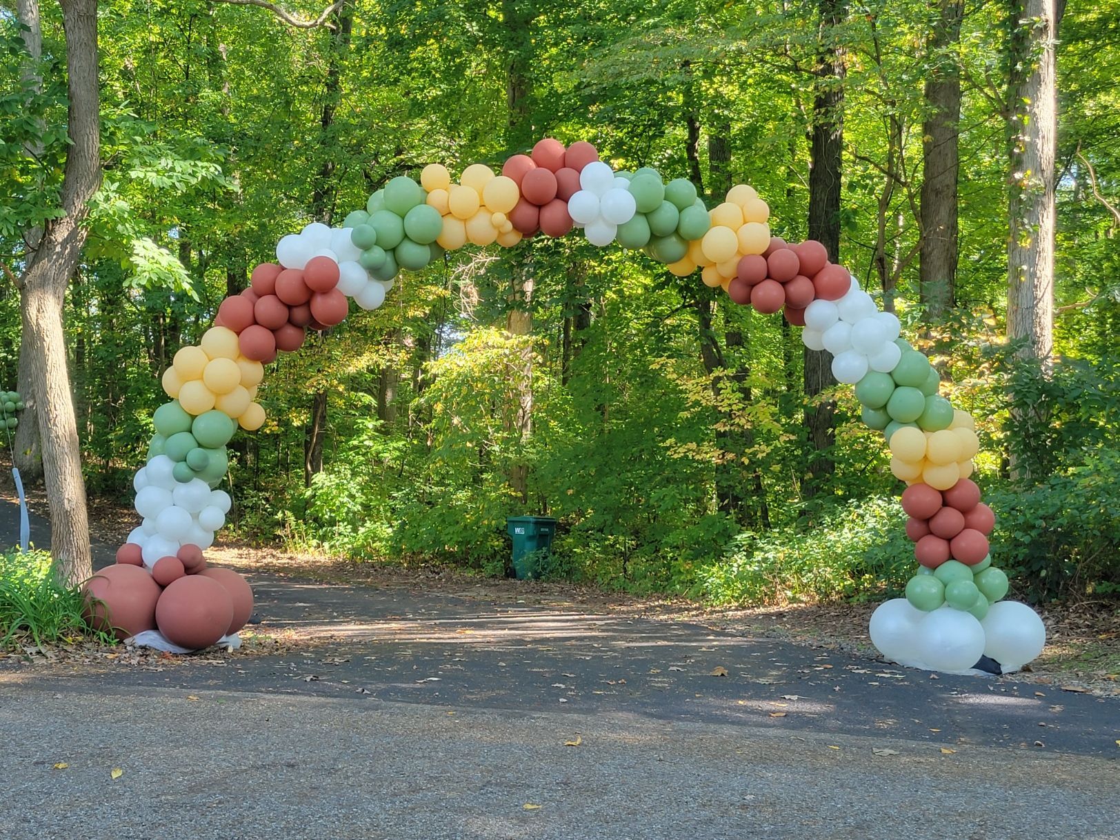 Balloon arch in fall colors over a paved path in a wooded area.