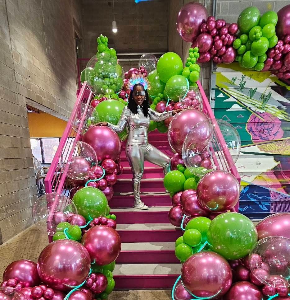 Person on staircase decorated with pink and green balloons, smiling, and wearing a silver suit.