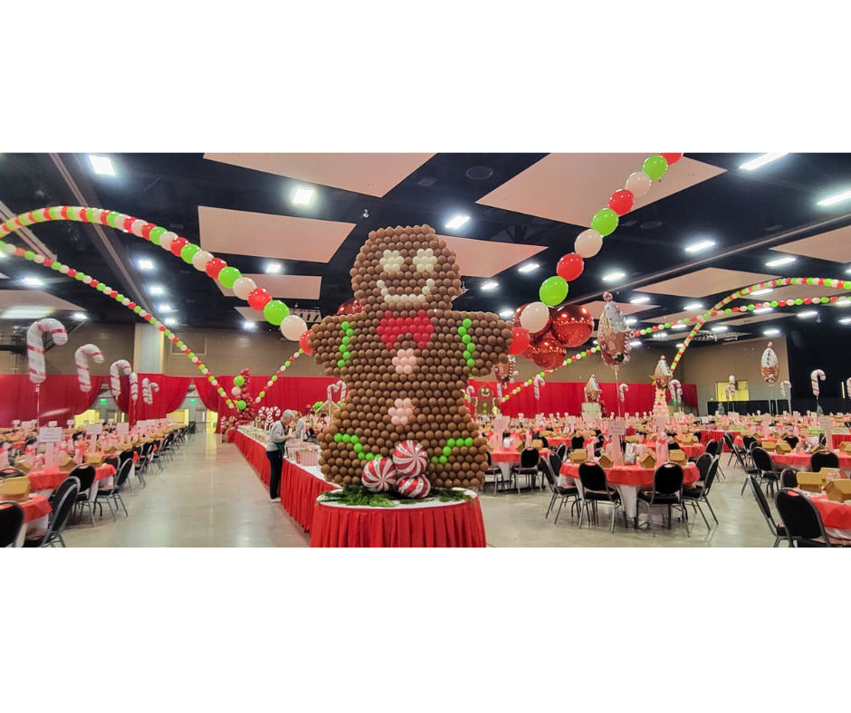 A large gingerbread man balloon sculpture at a holiday event. Tables and festive decorations fill the room.