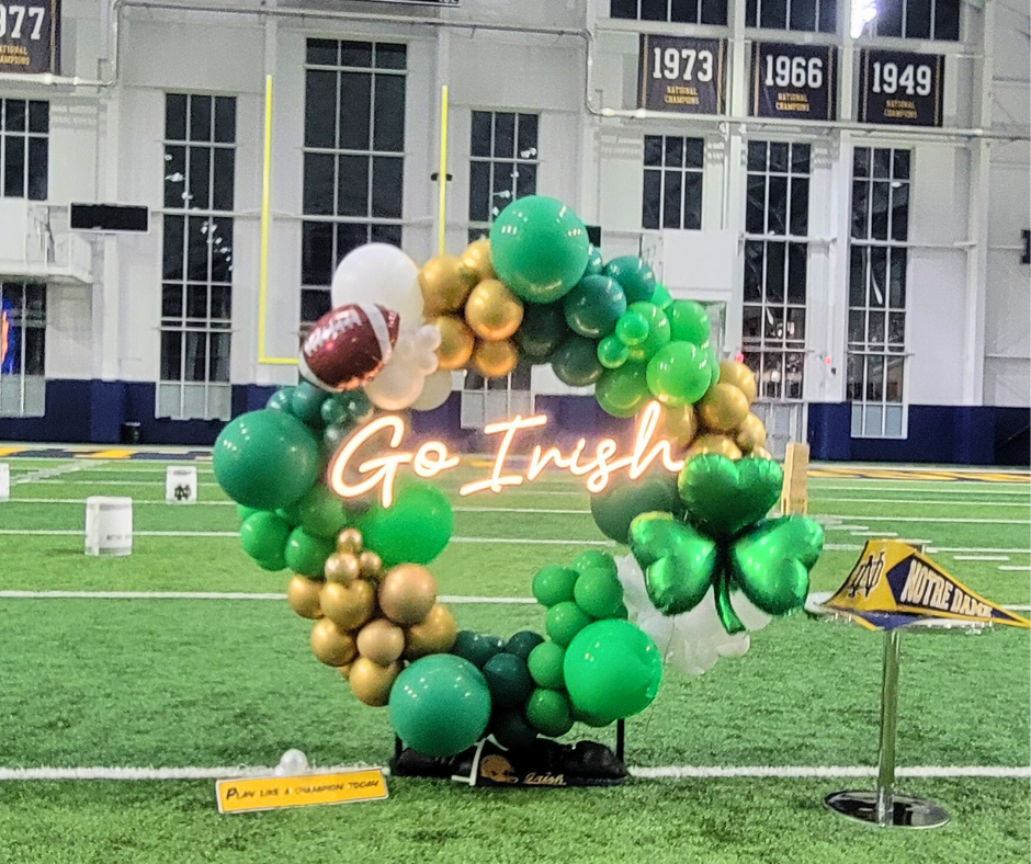 A wreath of green and gold balloons on a football field with a neon sign that says `` go irish ''.
