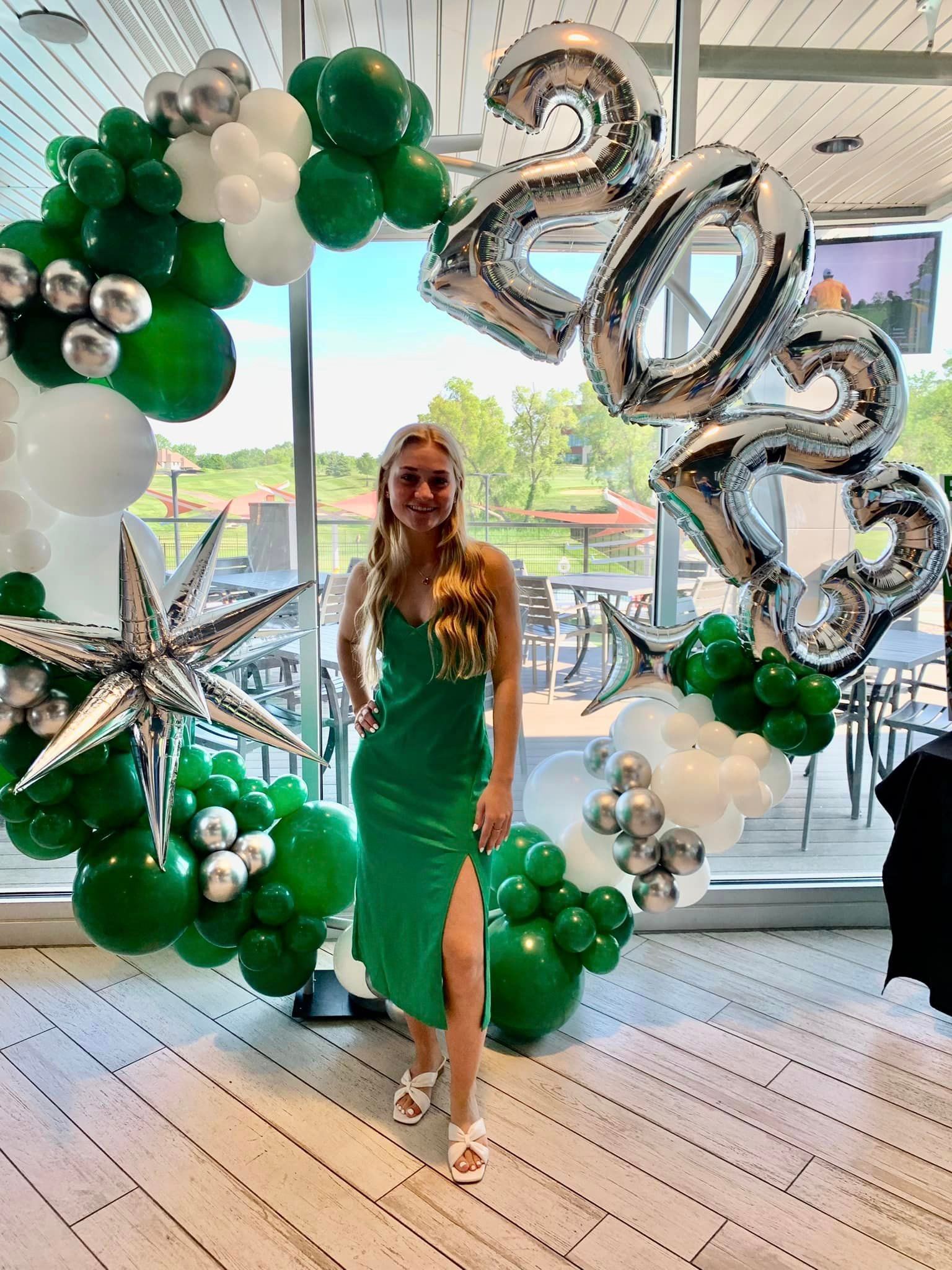 A woman in a green dress is standing in front of a balloon arch.