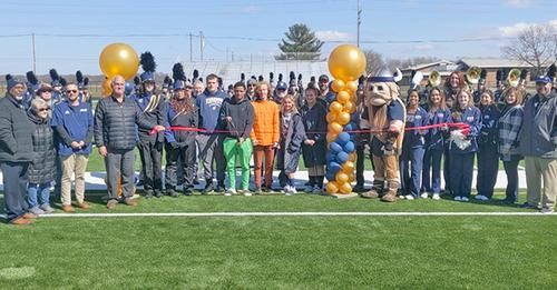 People cutting a ribbon on a new turf field, with a mascot, balloons, and band members in the background.