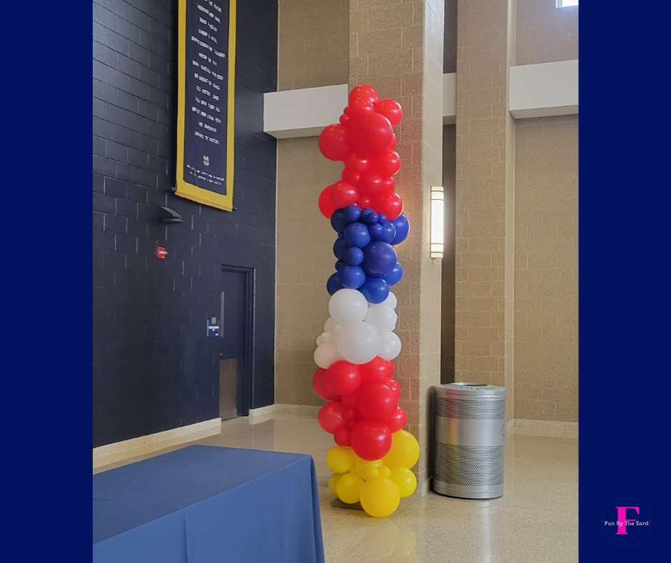 Column of red, white, blue, and yellow balloons in an indoor setting; near a blue table and a metallic trash can.