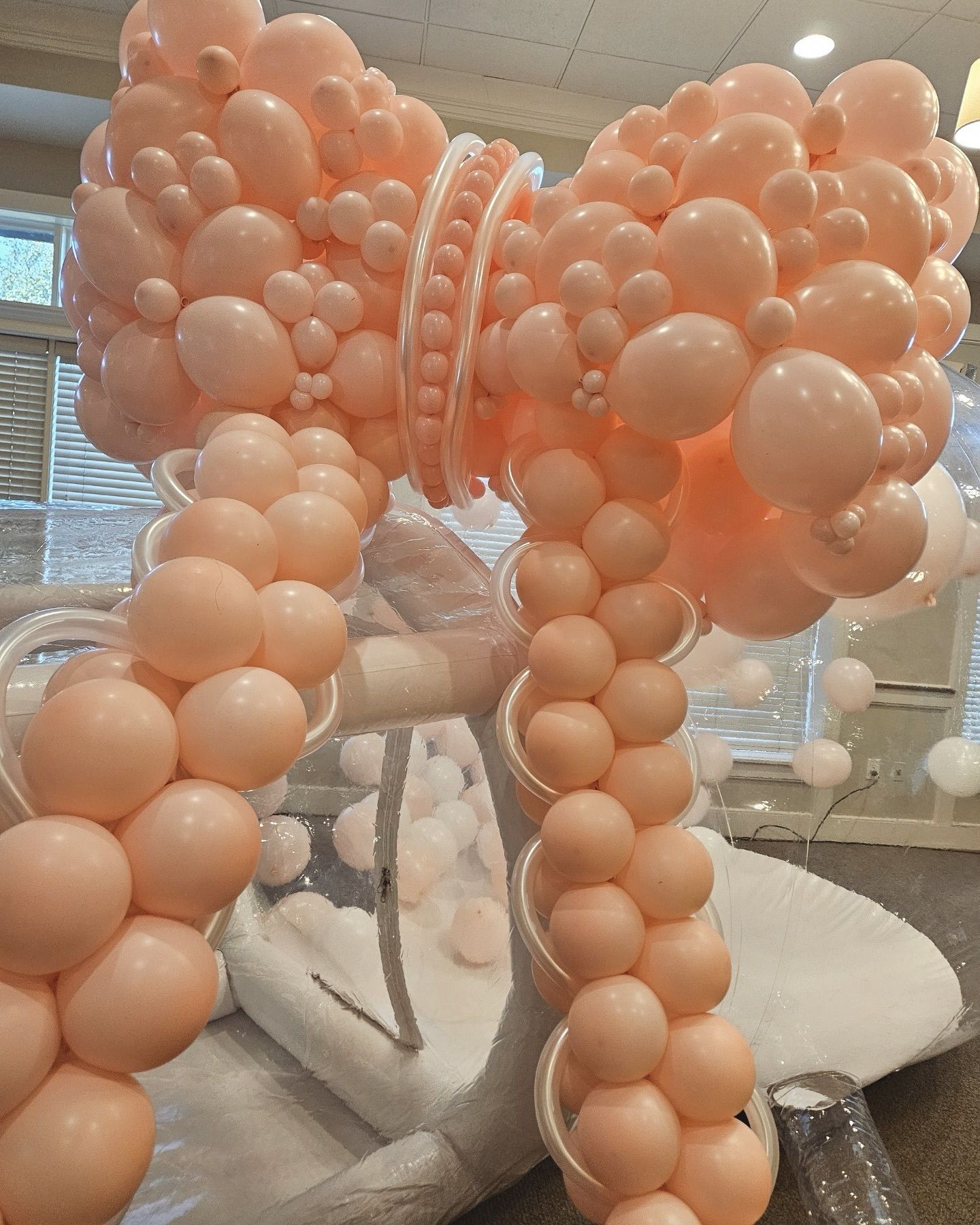 Large peach-colored balloon bow decoration, indoors, against a window and white decor.