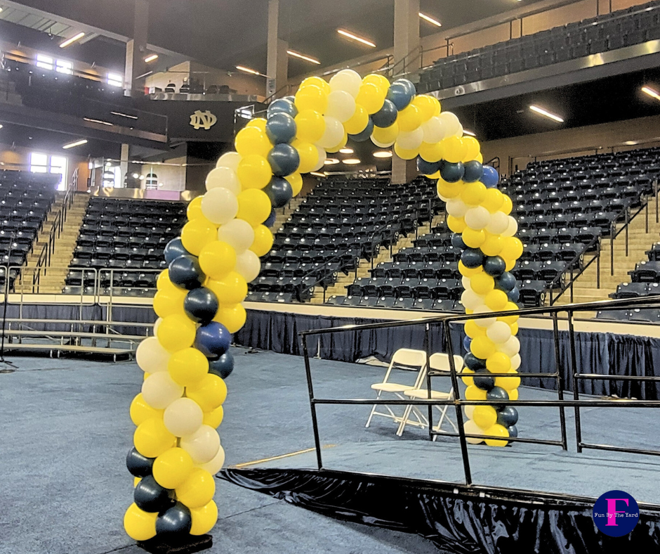 Balloon arch in yellow, navy, and white, inside an arena with rows of blue seats.