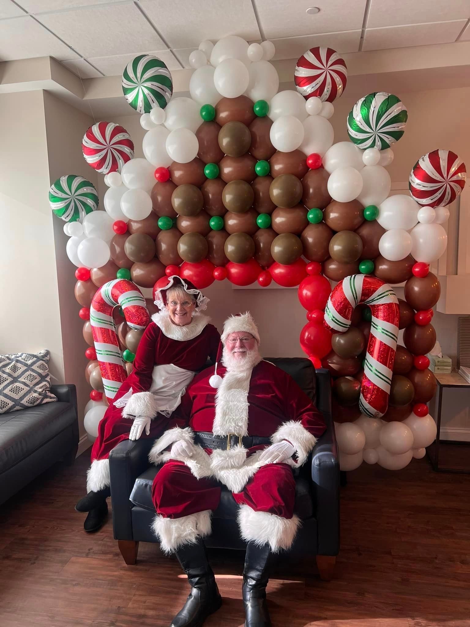 Santa Claus and Mrs. Claus seated in front of a gingerbread house made of balloons.
