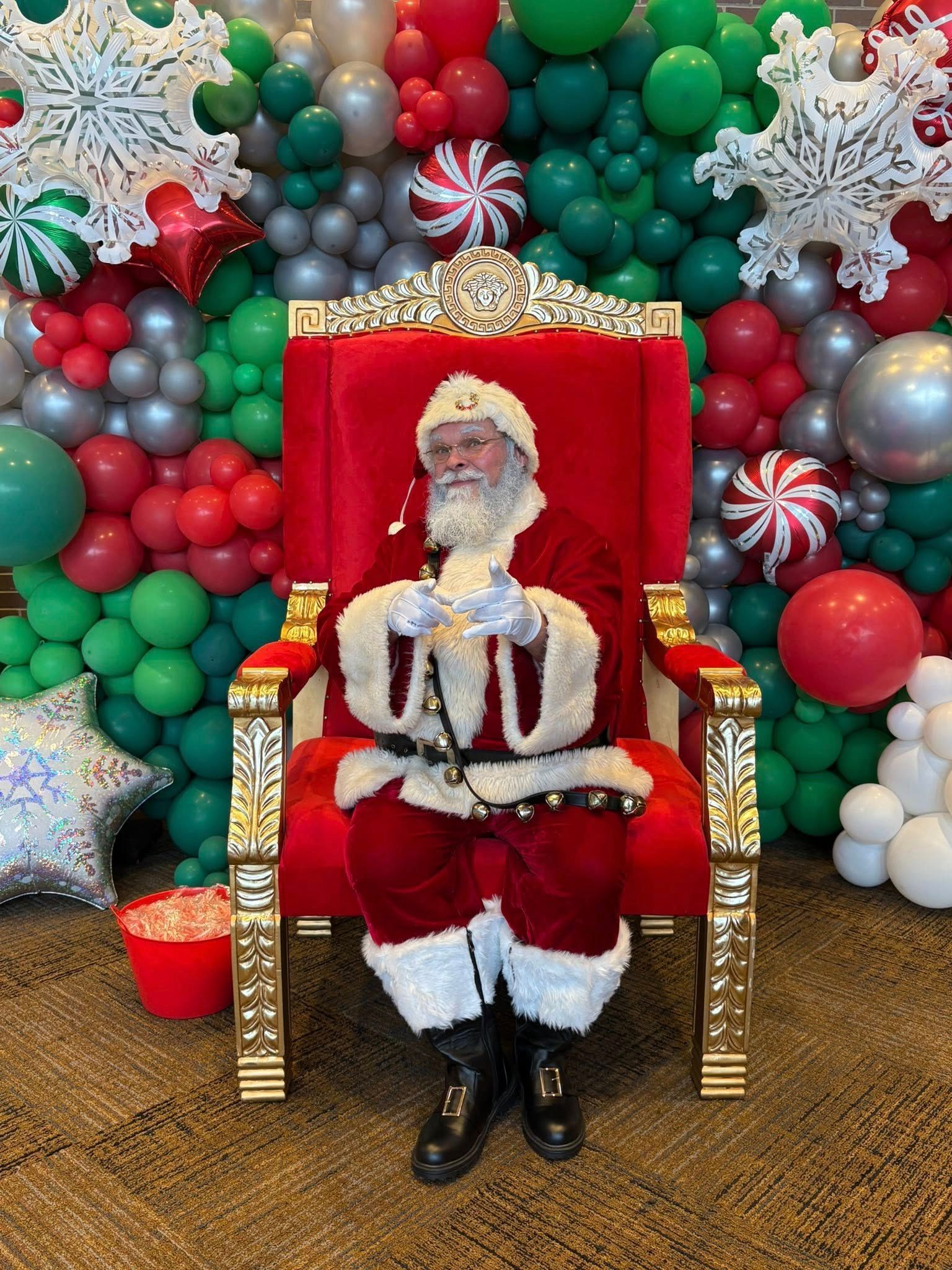 Santa Claus sits in a red throne before a balloon backdrop.