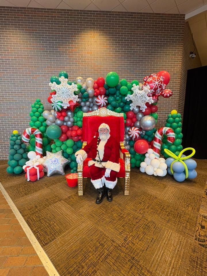 Santa Claus sits in a red throne before a festive balloon backdrop.