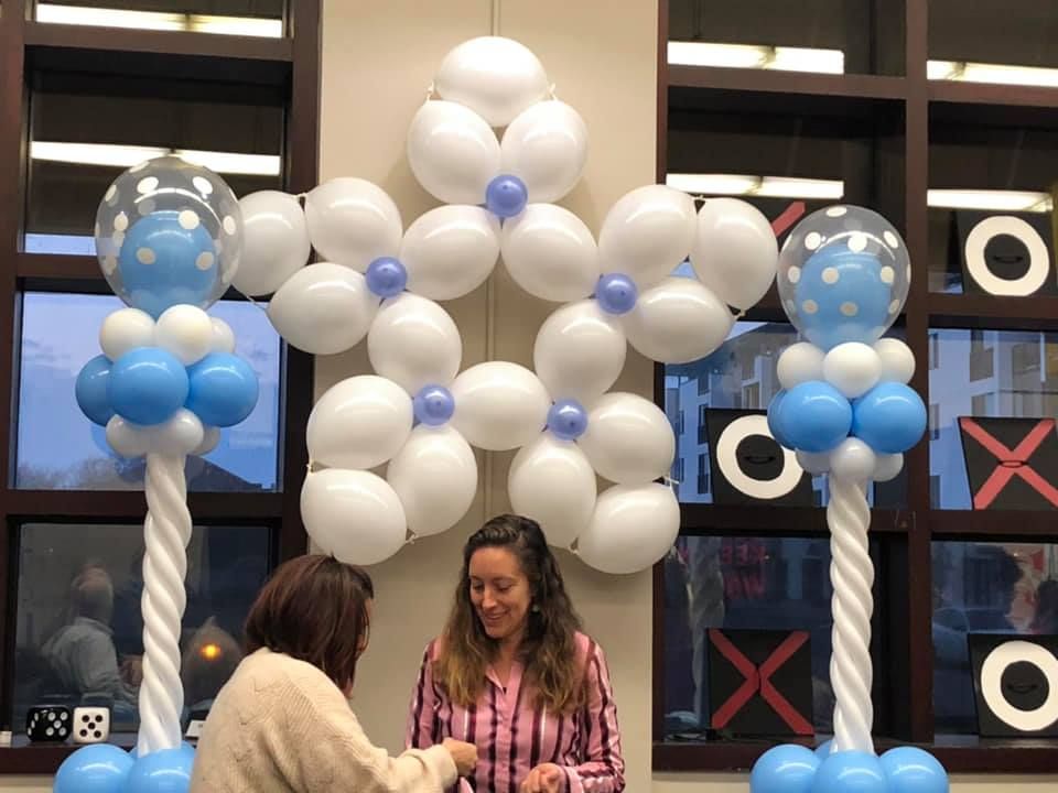 Two women are standing in front of a wall decorated with blue and white balloons.