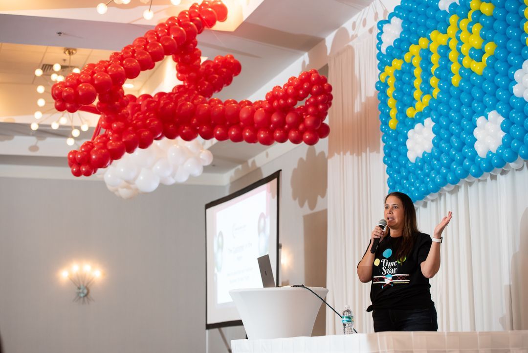 A woman stands at a podium with balloons in the shape of an airplane hanging from the ceiling