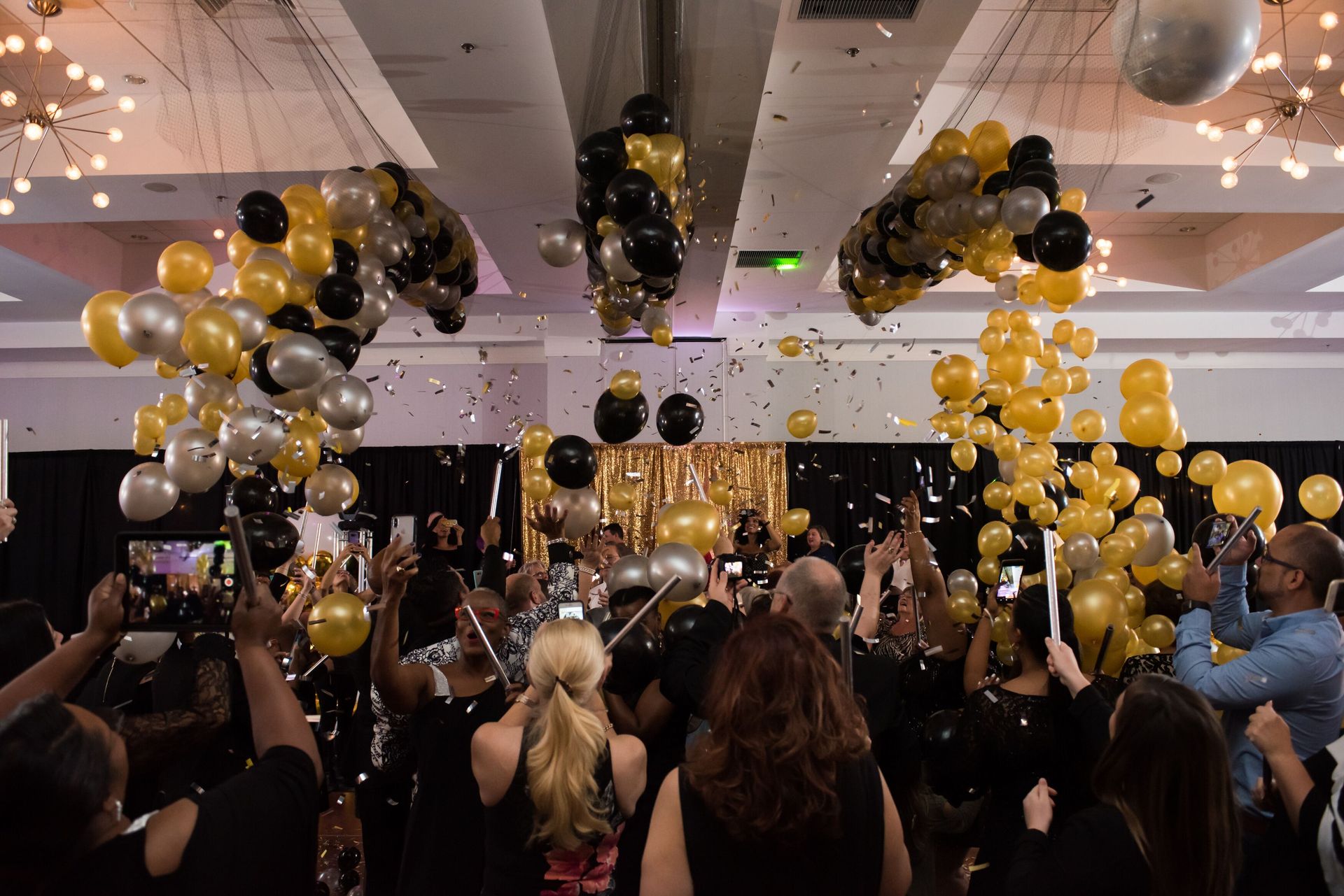 A crowd of people are standing in a room with balloons and confetti falling from the ceiling.  Balloon Drop