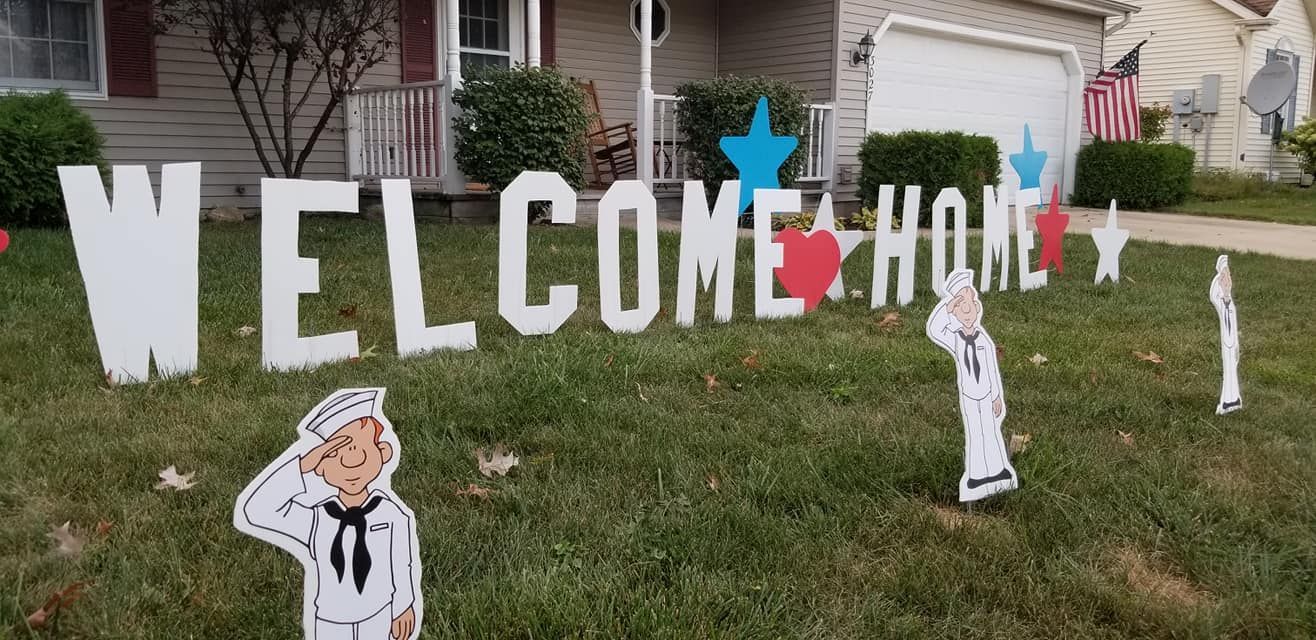 A welcome home sign is in front of a house.  Navy themed yard card