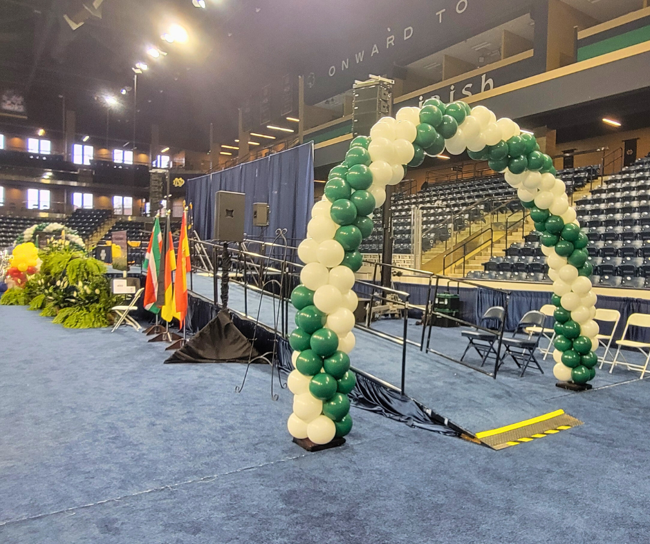 Green and white balloon archway at an indoor event, with chairs and banners in the background.