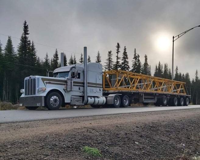 Un semi-remorque blanc transporte une charpente métallique jaune sur une route bordée d'arbres sous un ciel nuageux.