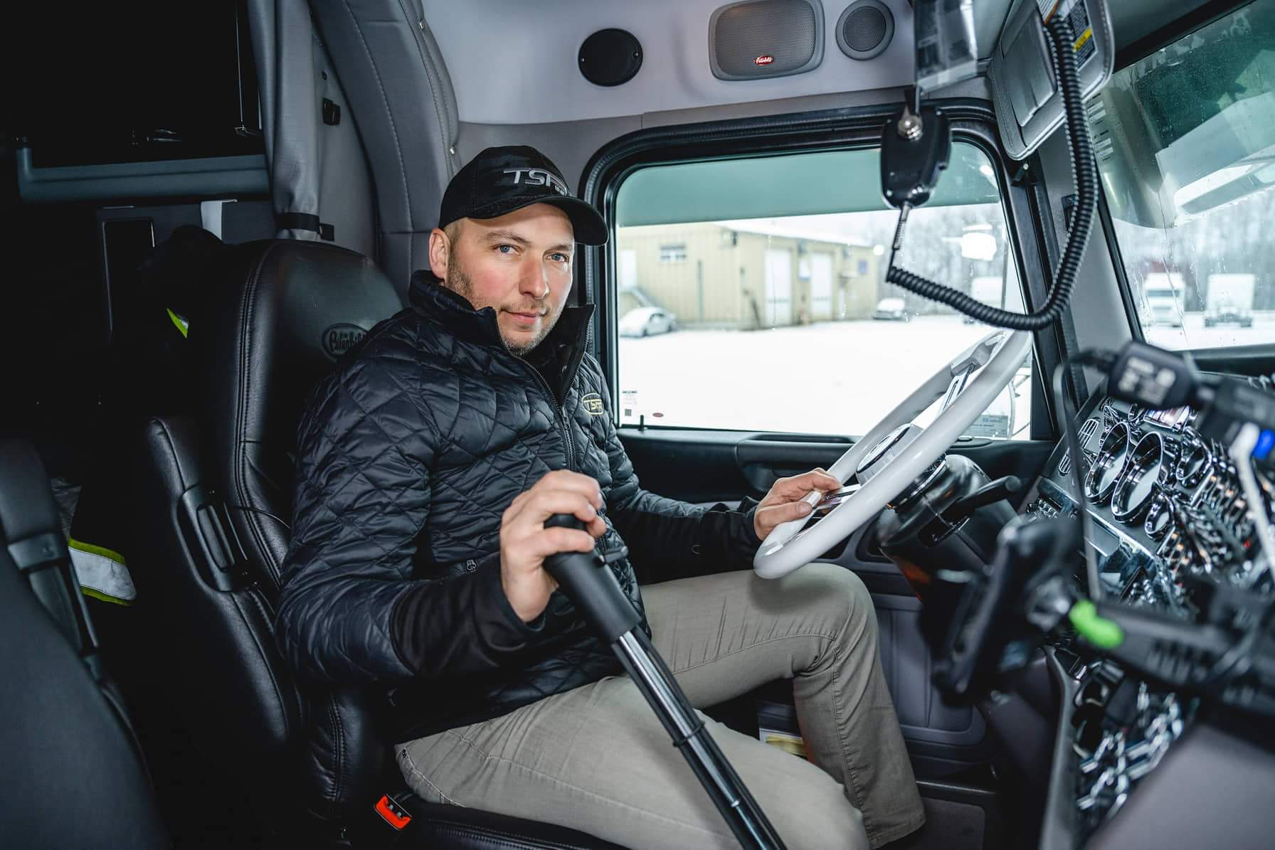 Un homme portant une veste noire et un chapeau est assis sur le siège conducteur d'un semi-remorque, tenant le levier de vitesse, regardant la caméra.
