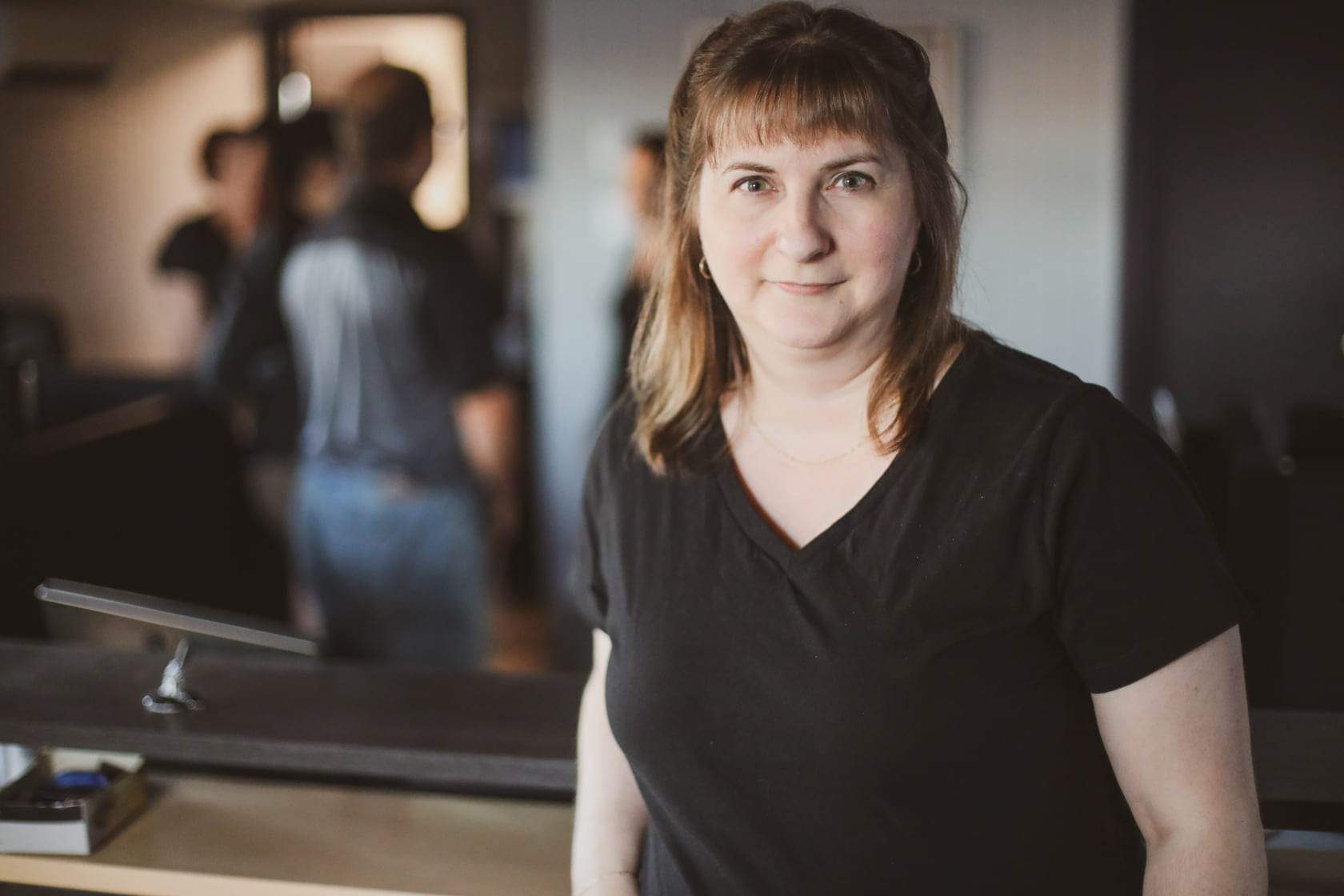 Woman with light brown hair and a black v-neck shirt smiling in an office setting, with people blurred in the background.