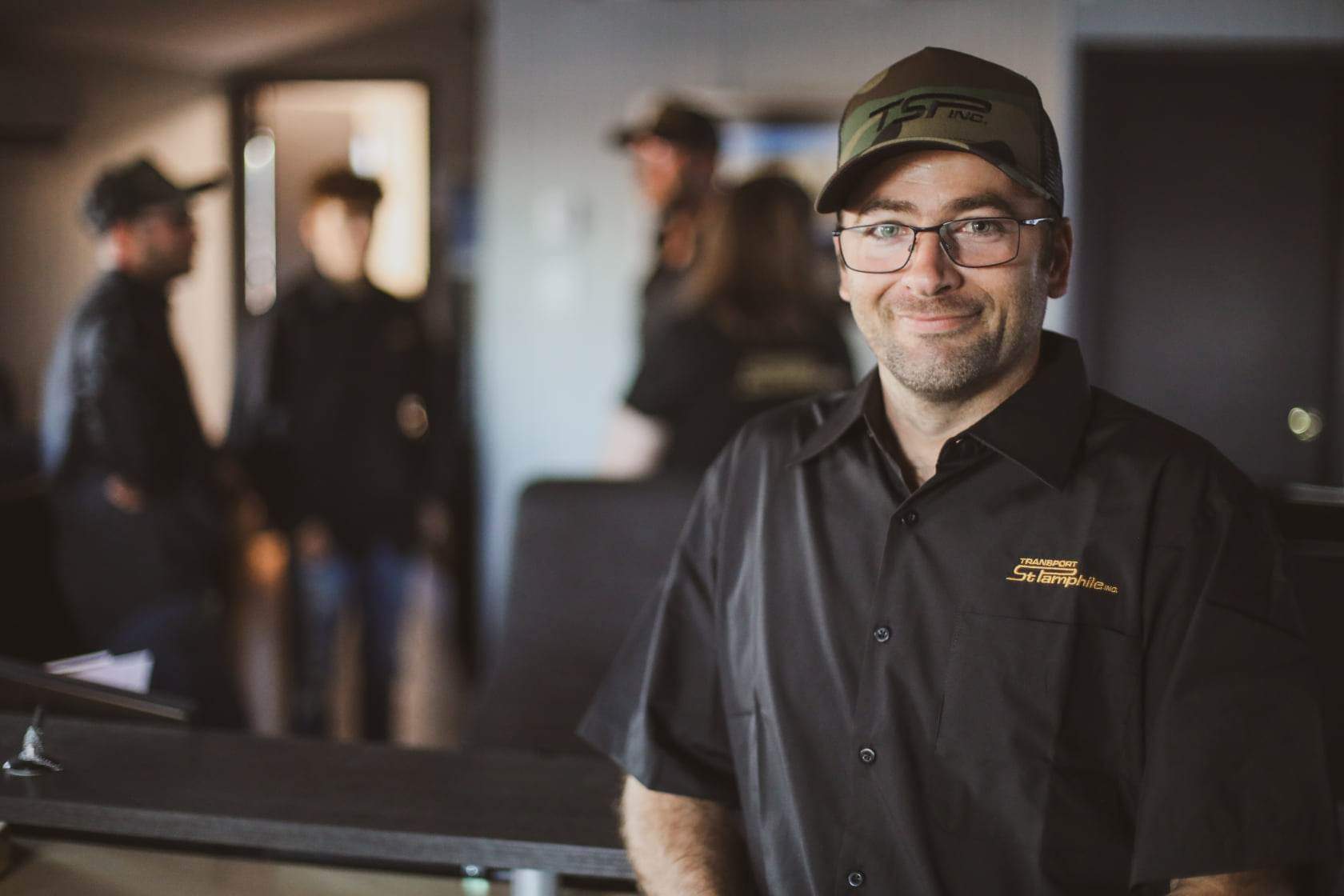 Man in a black work shirt and camo hat smiles at the camera; a group of people stand blurred in the background.