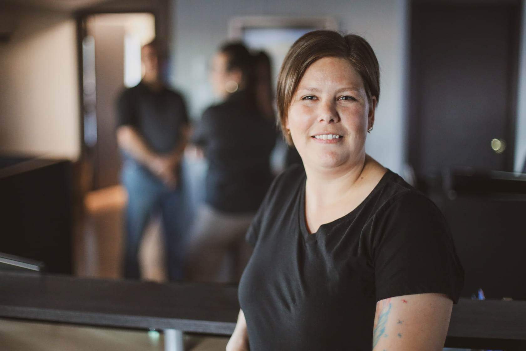 Woman with short brown hair smiles at the camera. She wears a black shirt and stands in an office with two blurred figures in the background.