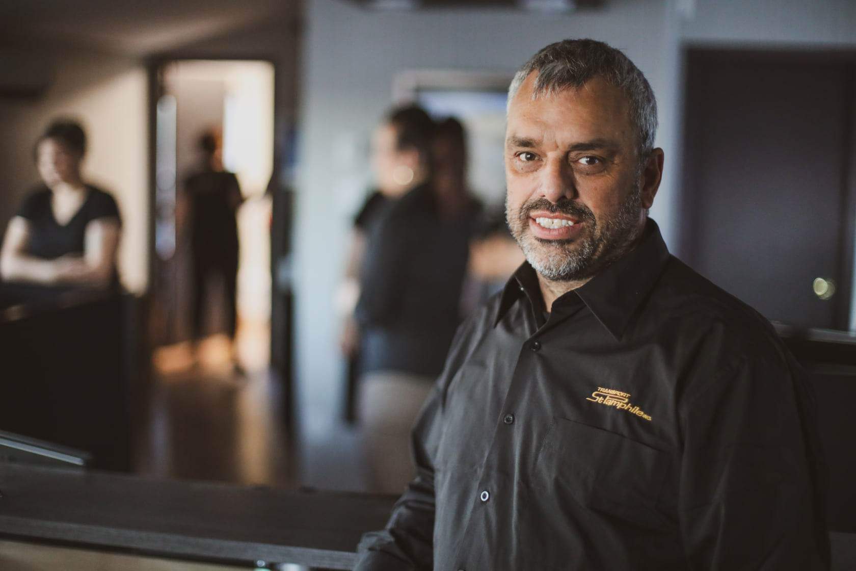 Man with graying beard smiles, standing behind a counter in a dimly lit room. Other people blur in the background.