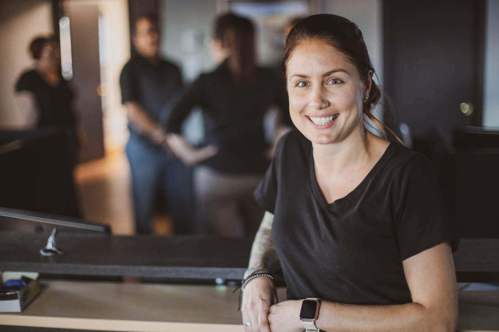 Woman with a tattoo smiles at the camera while leaning on a counter. Three coworkers are blurred in the background.