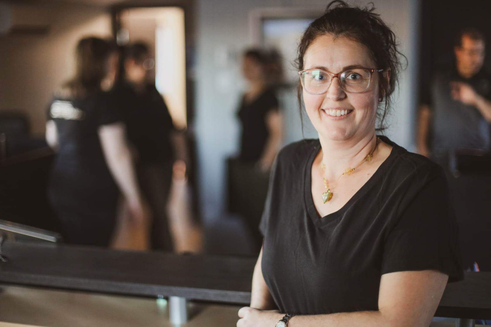 Woman with glasses smiles at the camera, wearing a black shirt. Several blurred figures stand in the background.