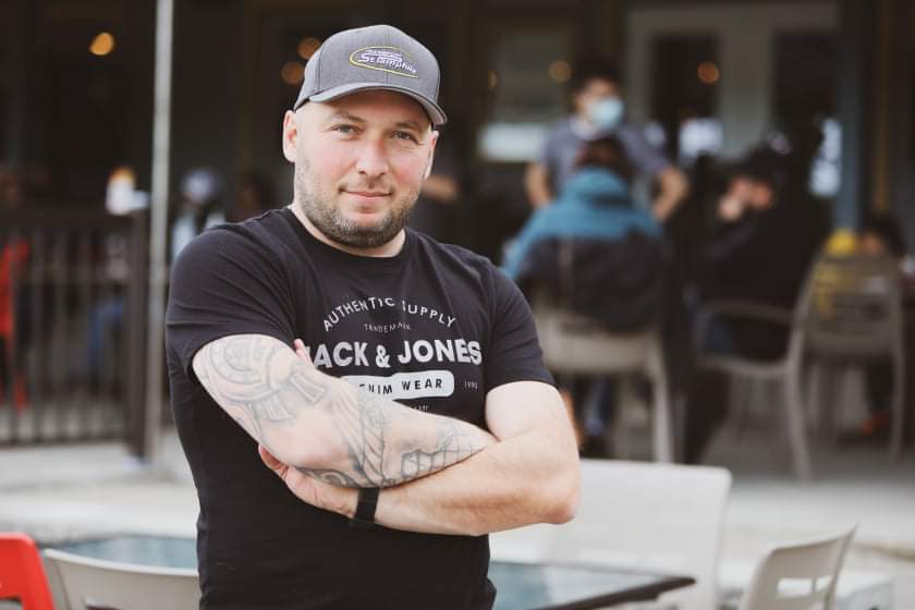 Man with crossed arms, wearing a baseball cap and t-shirt, outside a restaurant. He's smiling, with a tattoo visible on his arm.
