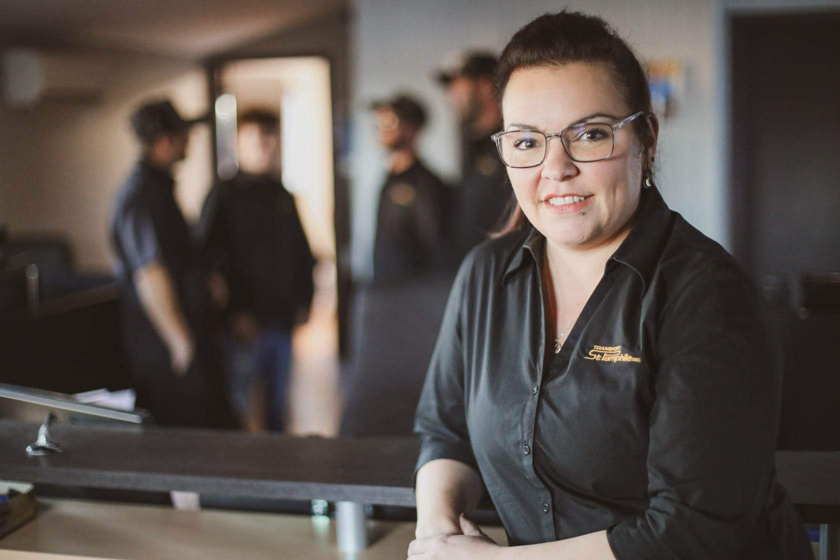 Woman in black shirt with glasses smiles at the camera, standing behind a counter. Several men in uniform are blurred in the background.