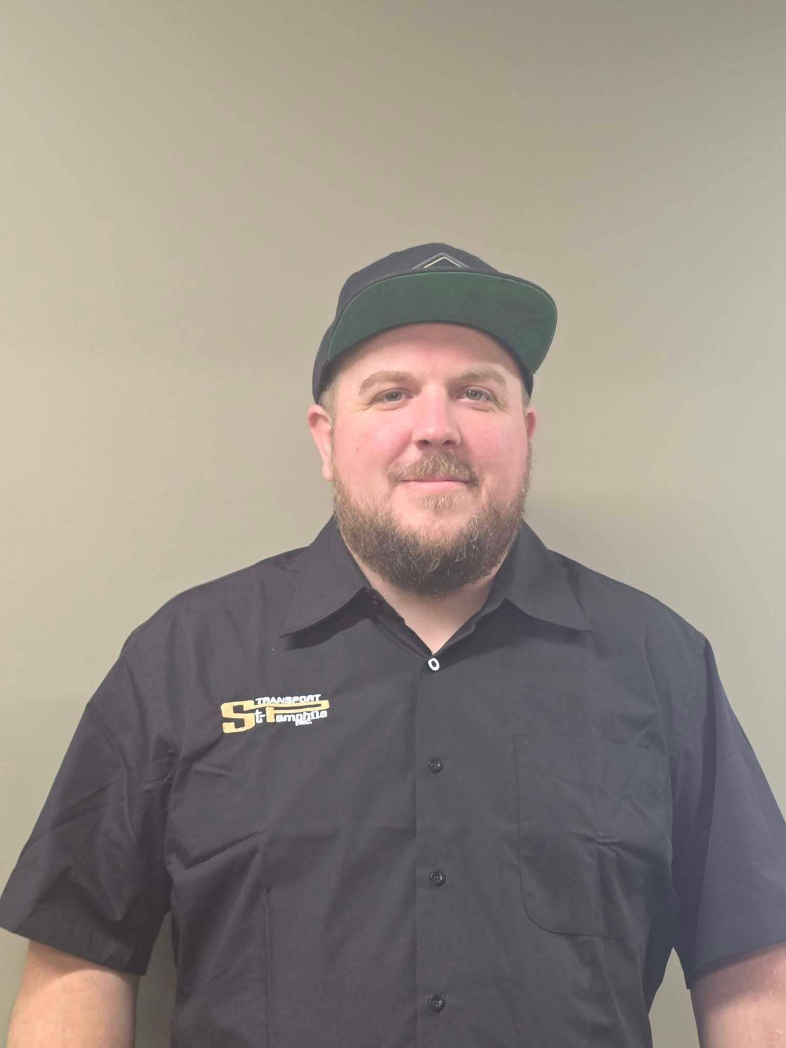 Man wearing a black hat and shirt with a company logo, against a plain wall.