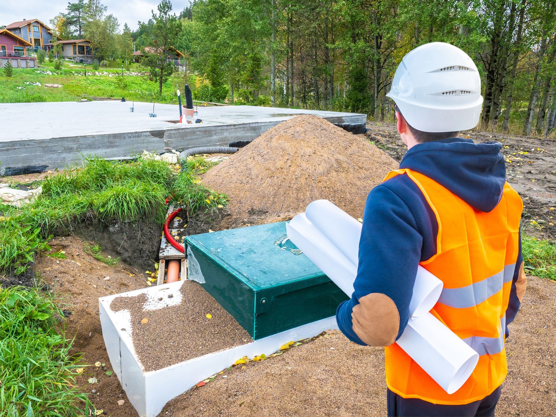 A man in an orange vest and hard hat is holding a blueprint in front of a construction site.