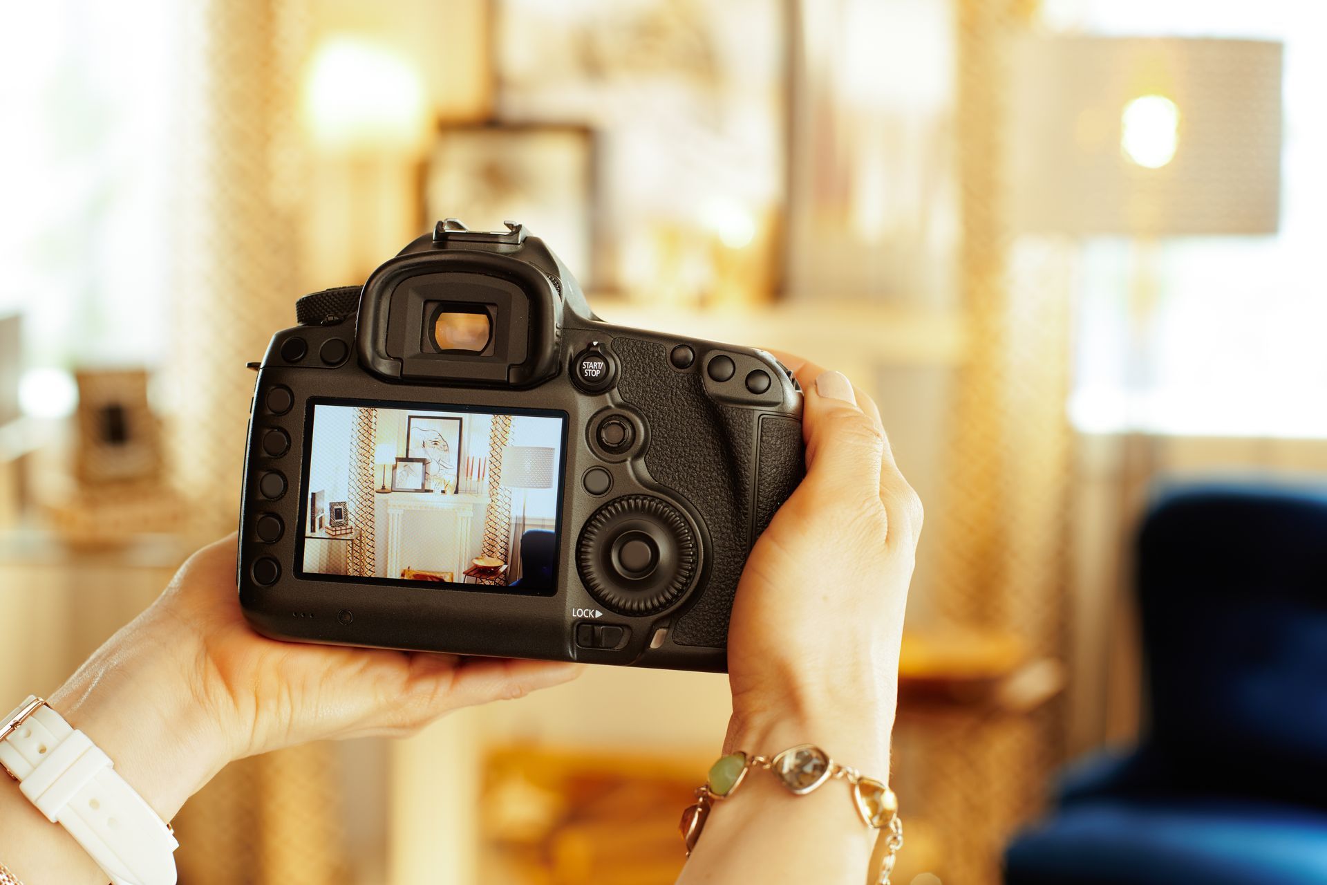Hands holding a digital camera, photographing an interior with warm lighting and a blue chair.