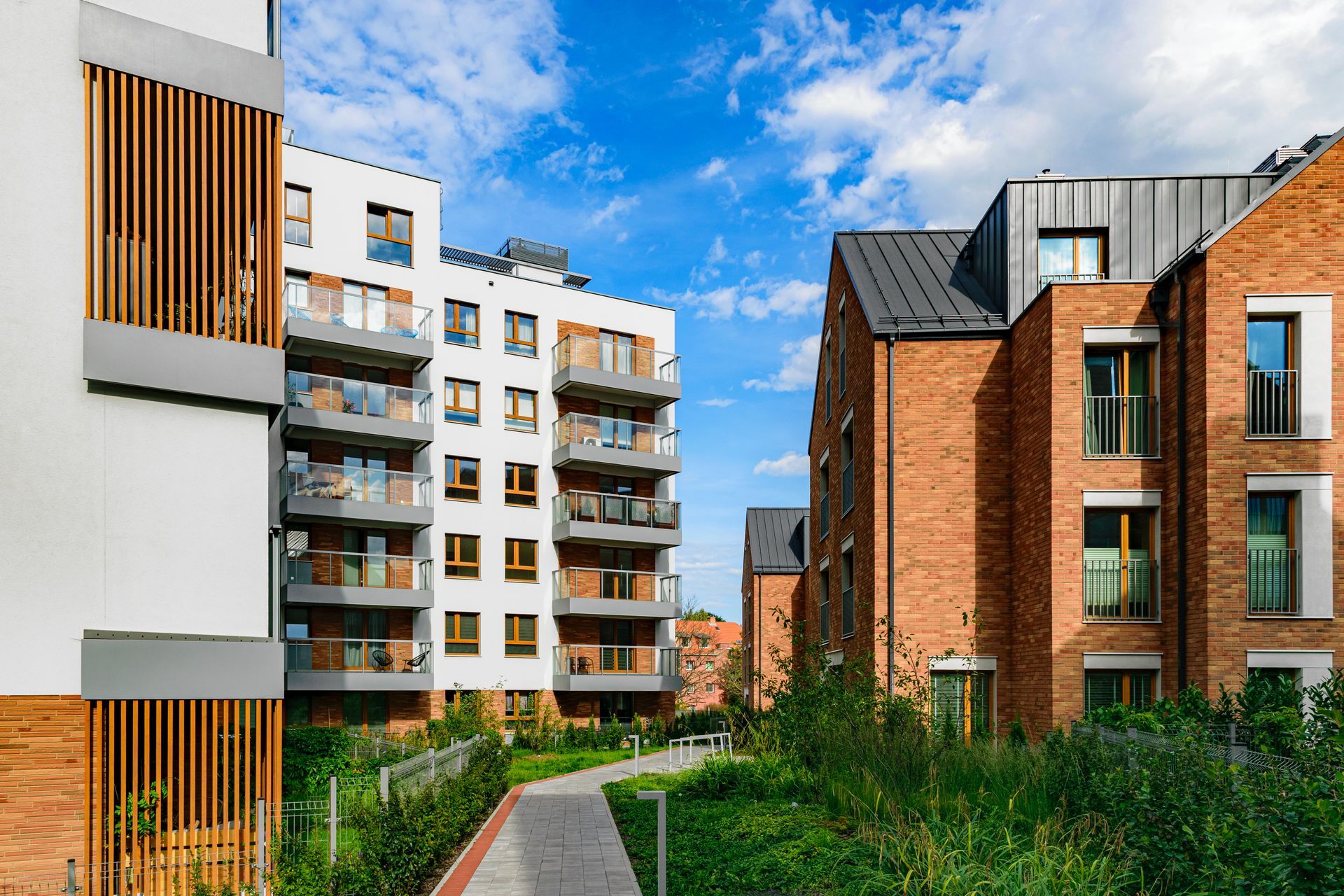Modern apartment buildings with brick and white facades under a blue sky, pathway through green grass.