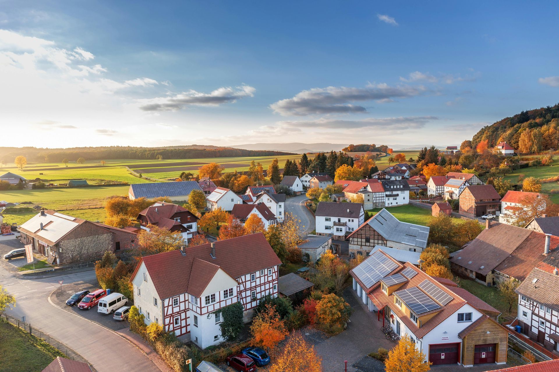 Aerial view of a small European village with red-roofed houses and autumn foliage under a blue sky.