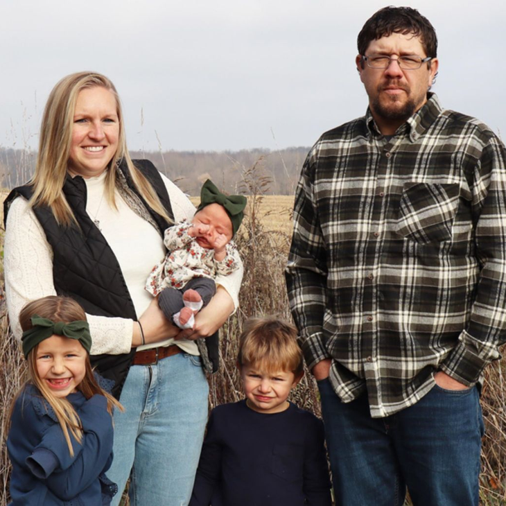 Family of five smiling outdoors. Mother holds baby, two children stand nearby, father smiles.